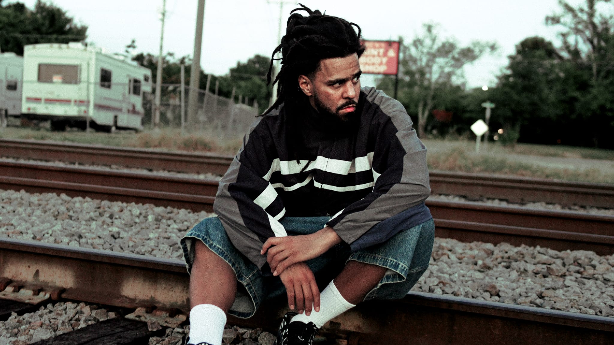J. Cole with dreadlocks sits on railroad tracks, wearing a striped jacket and denim shorts, looking contemplative.