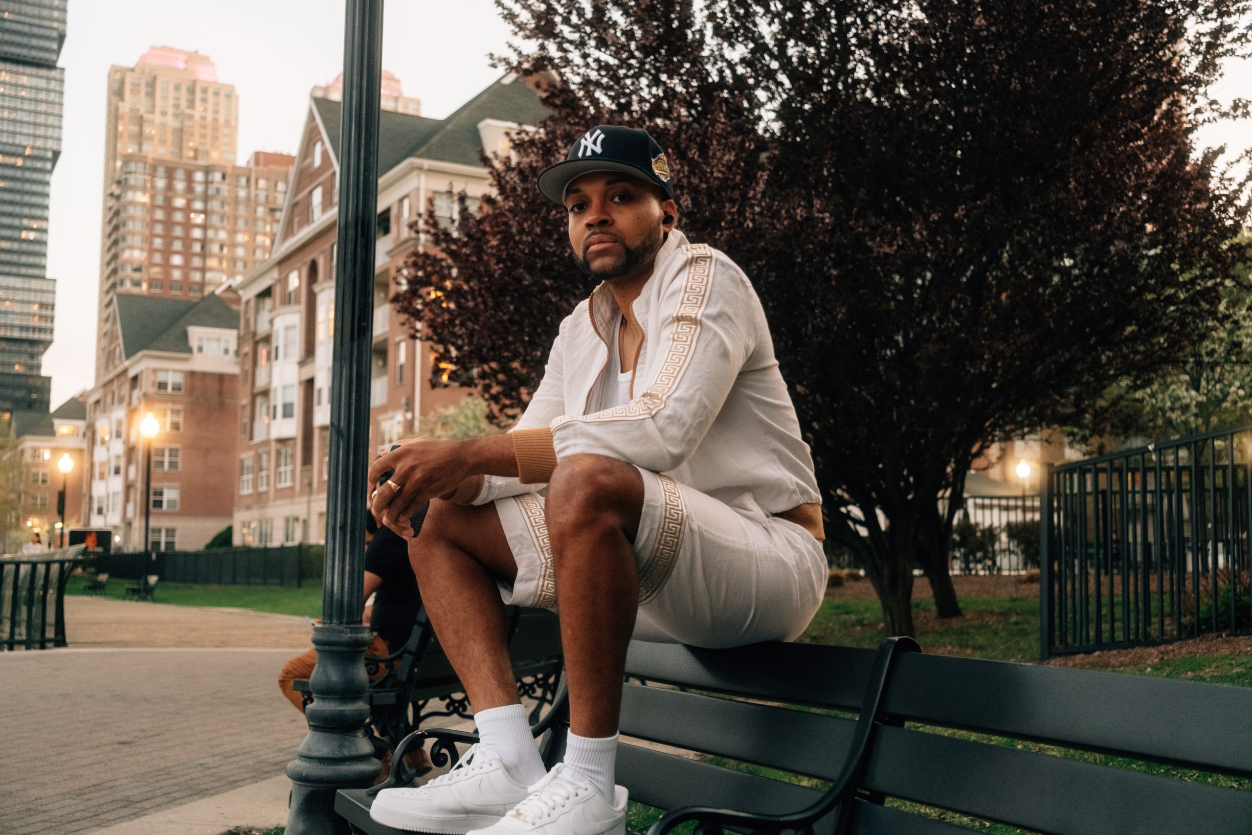 Cristo From The Bronx wearing a white outfit and New York Yankees cap while seated on a park bench.